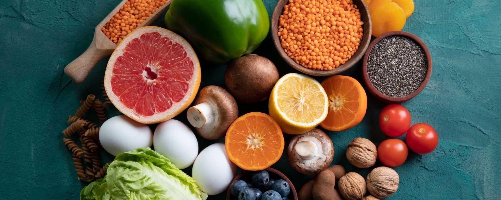 Una selección de alimentos antiinflamatorios naturales como frutas, verduras, nueces y semillas. Pomelo, limón, naranja, pimiento verde, champiñones, tomates cherry, nueces, arándanos, lechuga, huevos blancos, pasta rotini, orejones de albaricoque, lentejas rojas, semillas de chía.